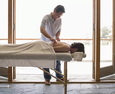 A massage therapist massages a client who is lying prone on a massage table. The doors to the patio are open behind them.