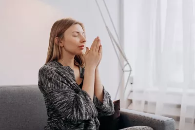 A woman sitting on a couch meditates with her hands together.