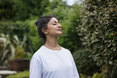 A woman with brown hair in a plain sleeved shirt takes a big deep breath with her eyes closed.