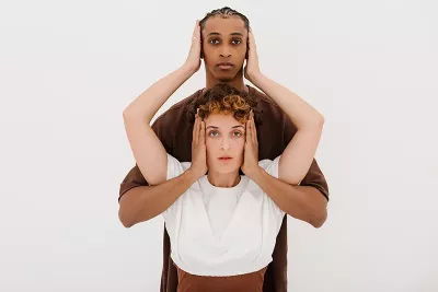 a taller Black person and a shorter White person cover the other's ears with two hands.