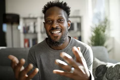 A Black man smiles and gestures largely with both hands.