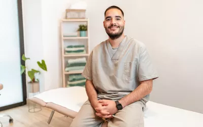 A massage therapist sits on the edge of a massage table, smiling for a photo.