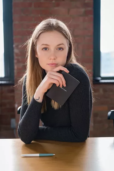 A woman holds a notepad and stares directly into the camera while sitting at a wood table.