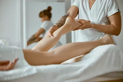A massage therapist dressed in white works on a prone client's lower calf.