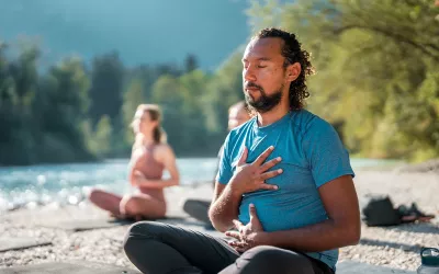 A man and two women have their hands over their hearts while they practice breathwork on the shores of a lake.