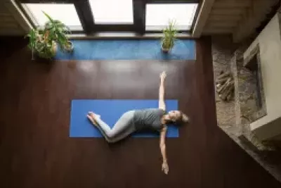 A woman lies on a blue yoga mat on the floor with her arms outstretched.