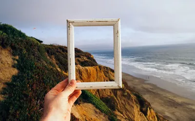 Hand holding small picture frame with ocean and cliffs in the background.