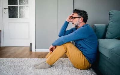 A man with dark hair and a beard sitting on a rug holding his head in his hands looking stressed.