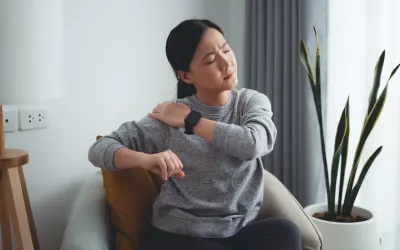 A woman sitting in a chair grabs her shoulder in pain.