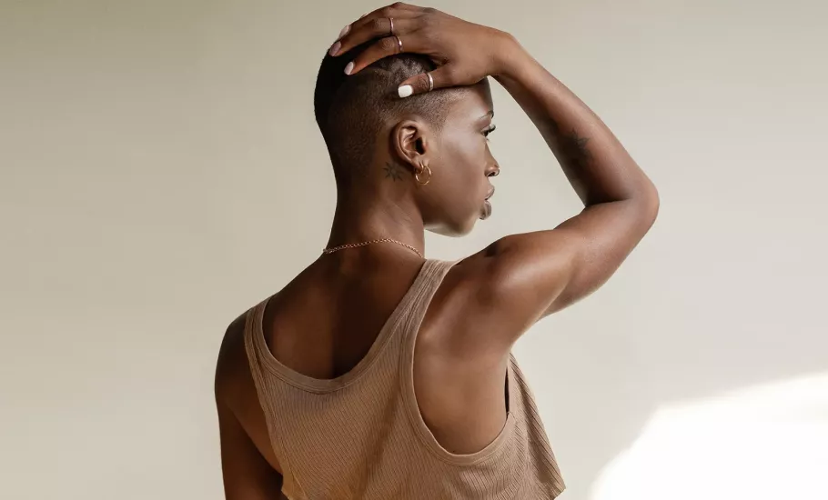 a Black woman in a tank top with a buzz cut holds her arm over her head, rotating her scapula.