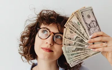 Woman with curly hair and glasses holding fan of money and looking up