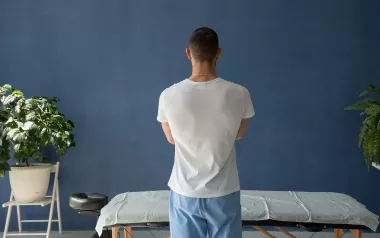 A massage therapist stands over a massage table, facing away from the camera in a pensive tone.