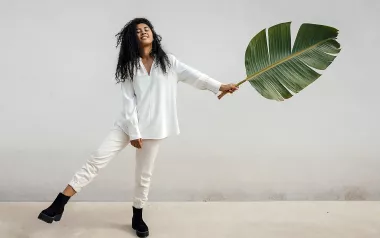 A woman balances on one leg while holding a giant leaf in the other hand, almost as a counterbalance to her raised leg.