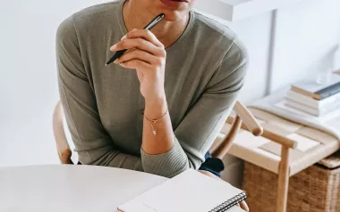 A woman in a long-sleeve shirt holds a black pen to her mouth as she thinks.