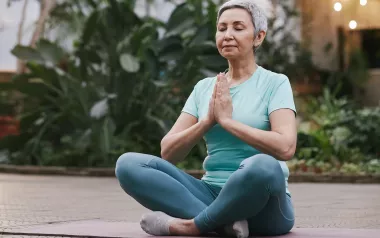 older woman with short hair seated criss-cross on a mat with her hands at heart center.