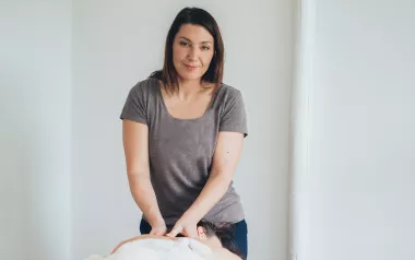 A female MT works on a prone client on a table. The MT is looking at the camera. 