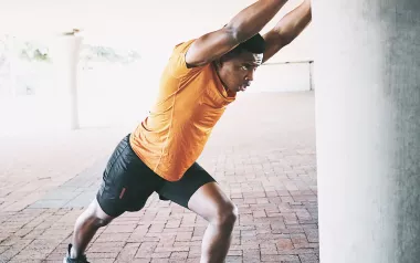 A Black man in exercise clothes pushes against a concrete column to stretch.