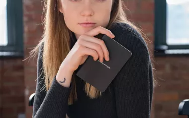 A woman holds a notepad and stares directly into the camera while sitting at a wood table.