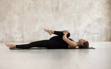 A woman on her back on a yoga mat stretches her right leg and glutes.