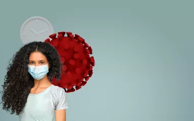 A woman with curly long hair wearing a face mask stands in front of a cartoon red cell.