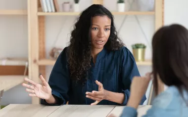 a woman with curly hair in a sweater sitting at a desk gestures with her hands.