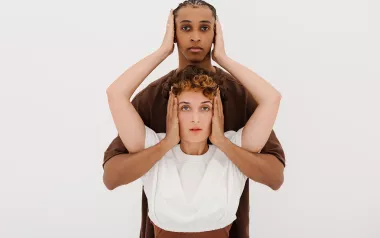 a taller Black person and a shorter White person cover the other's ears with two hands.
