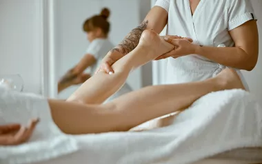 A massage therapist dressed in white works on a prone client's lower calf.