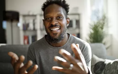 A Black man smiles and gestures largely with both hands.