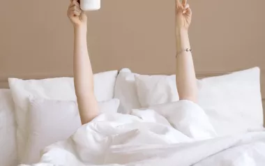 A person under the covers in bed holds up a peace sign with one hand and a coffee cup in the other hand.