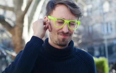 A man in a navy sewater and neon glasses clutches his ear and winces.