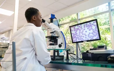 A young Black woman in a labcoat analyzing cells underneath a microscope.