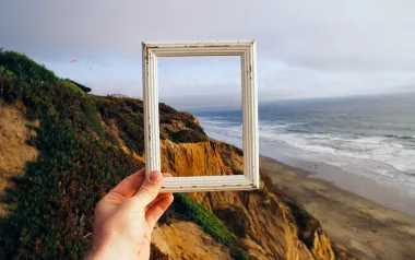 Hand holding small picture frame with ocean and cliffs in the background.