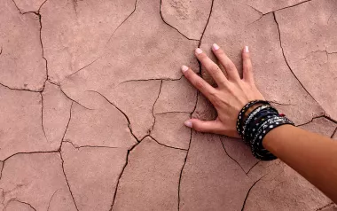 A hand with multiple bracelets touches cracked, dried dirt.