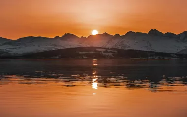 The sun sets over snowy peaks and a lake.
