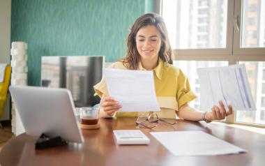 A woman smiles while looking at paperwork. A calculator and tablet are on the table in front of her.