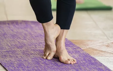 A pair of elderly feet stand on a yoga mat, one foot resting against the other.