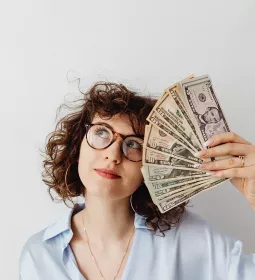 Woman with curly hair and glasses holding fan of money and looking up