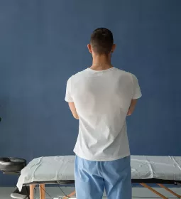 A massage therapist stands over a massage table, facing away from the camera in a pensive tone.