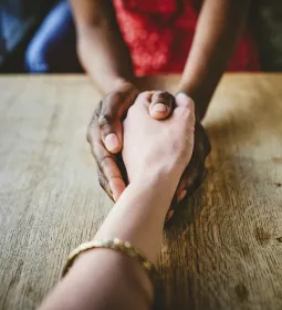 Two people grasping hands in a comforting manner across a table from each other.