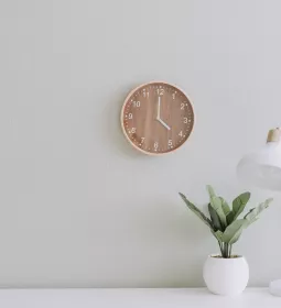 a wooden clock on a white wall next to a desk and plant.