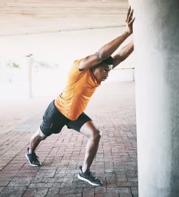 A Black man in exercise clothes pushes against a concrete column to stretch.