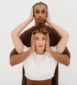 a taller Black person and a shorter White person cover the other's ears with two hands.