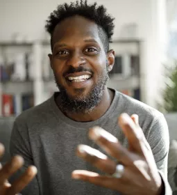 A Black man smiles and gestures largely with both hands.
