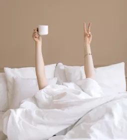A person under the covers in bed holds up a peace sign with one hand and a coffee cup in the other hand.