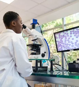 A young Black woman in a labcoat analyzing cells underneath a microscope.