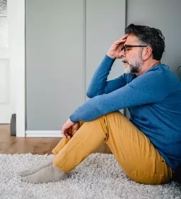 A man with dark hair and a beard sitting on a rug holding his head in his hands looking stressed.