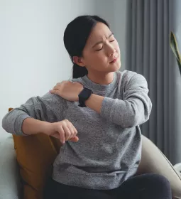 A woman sitting in a chair grabs her shoulder in pain.