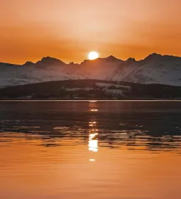 The sun sets over snowy peaks and a lake.