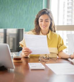 A woman smiles while looking at paperwork. A calculator and tablet are on the table in front of her.