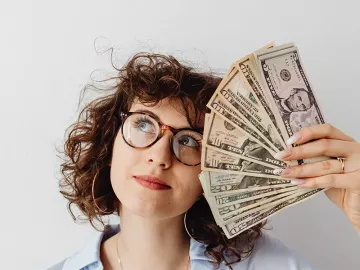 Woman with curly hair and glasses holding fan of money and looking up